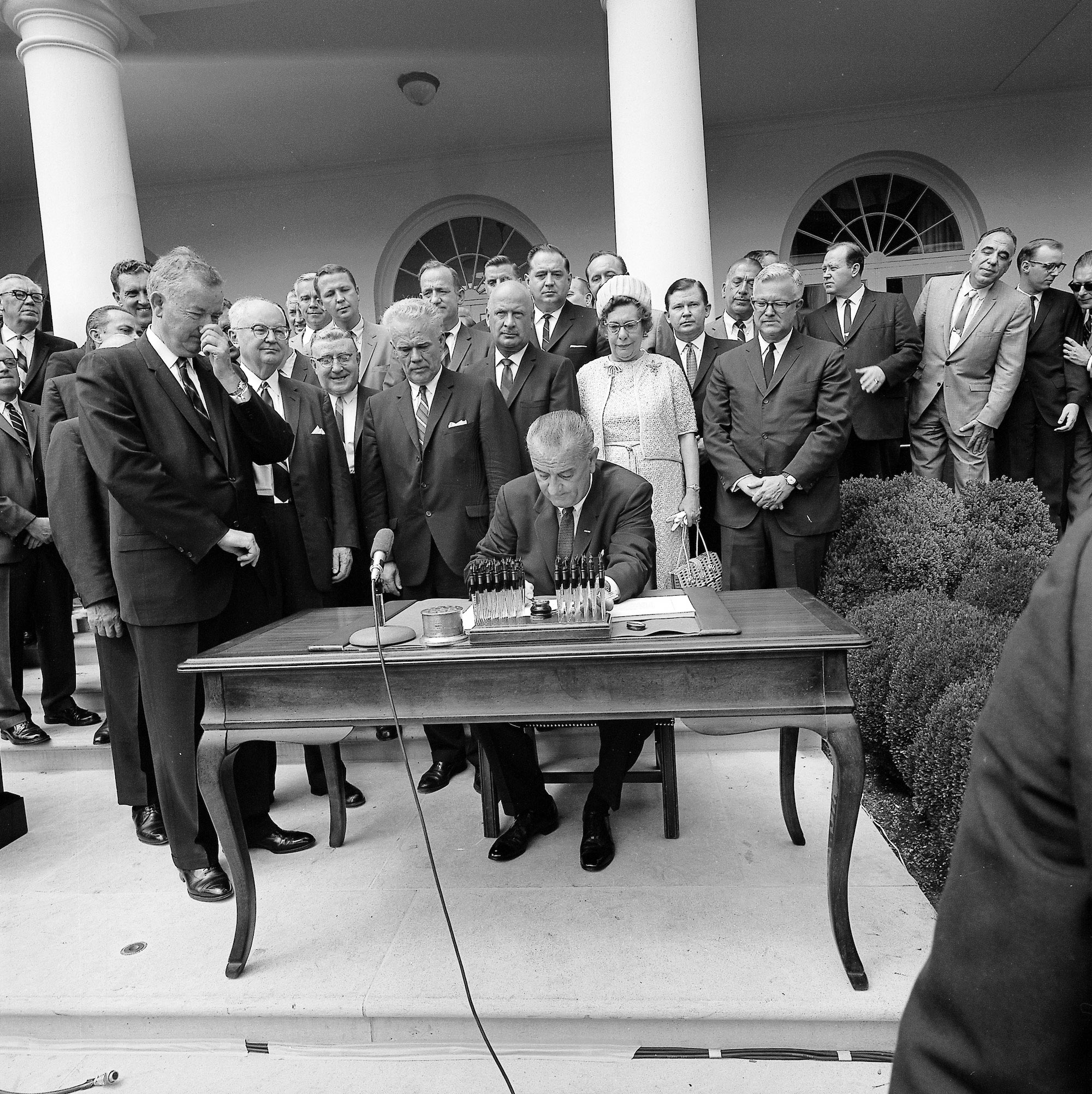 President Lyndon B. Johnson, seated at a table, signs the Housing and Urban Development Act as numerous guests look on.