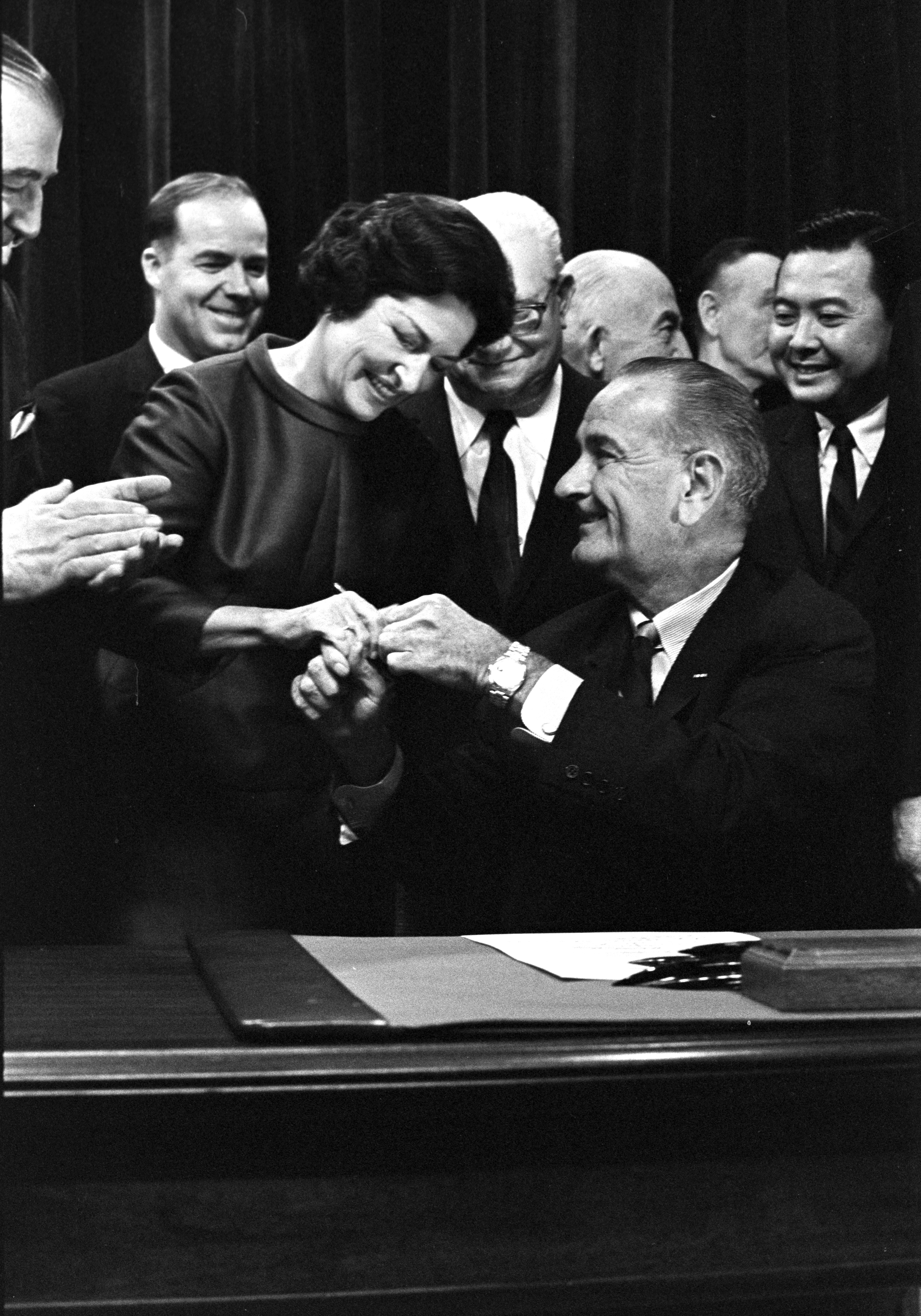 President Lyndon B. Johnson hands a signing pen to Lady Bird Johnson as others look on.