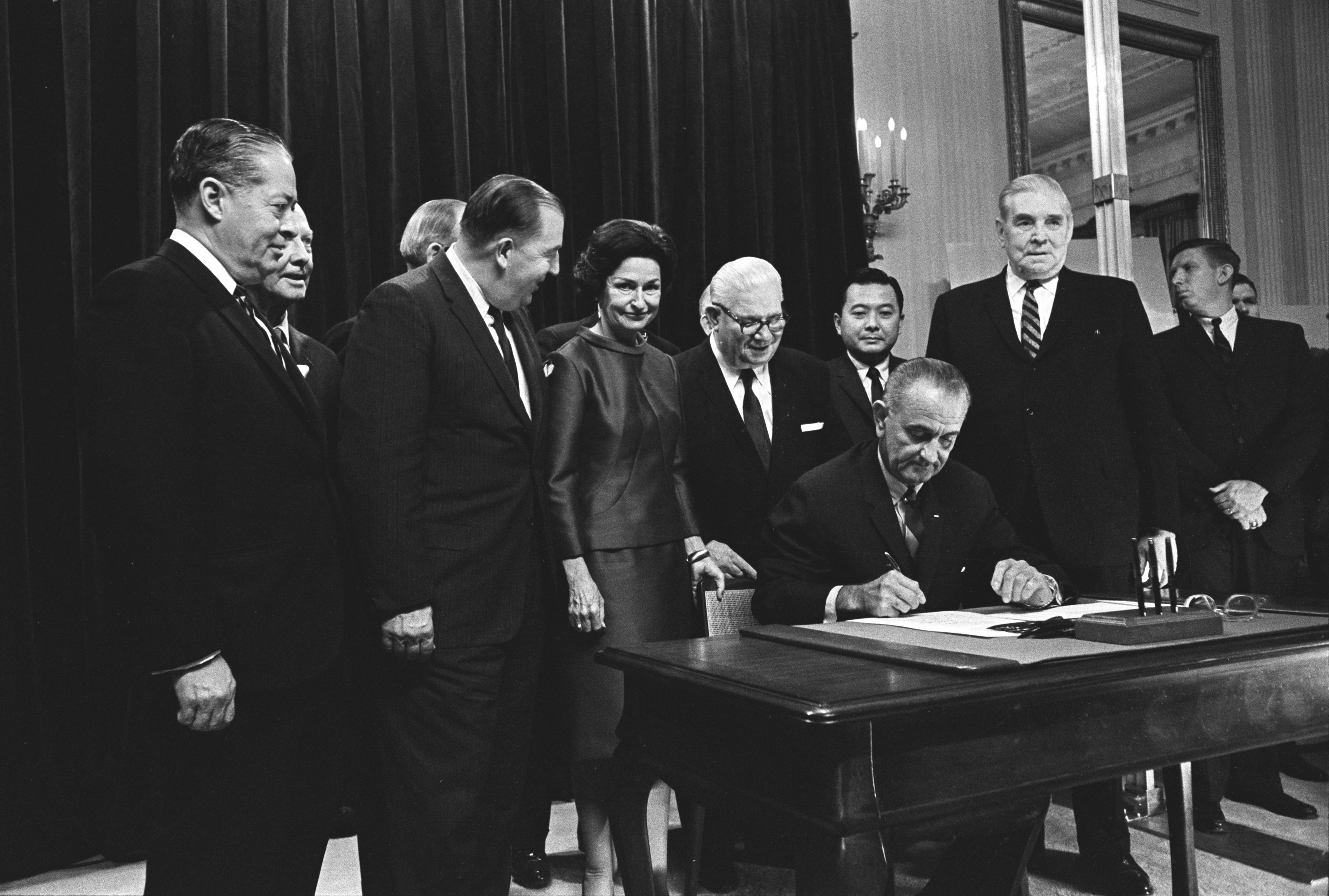 President Lyndon B. Johnson signing the Highway Beautification Act as others, including Lady Bird Johnson, look on. Sen. Jennings Randolph is at Mrs. Johnson's right. Sen. Daniel Inouye is behind LBJ, Sen. Pat McNamara is at his left.