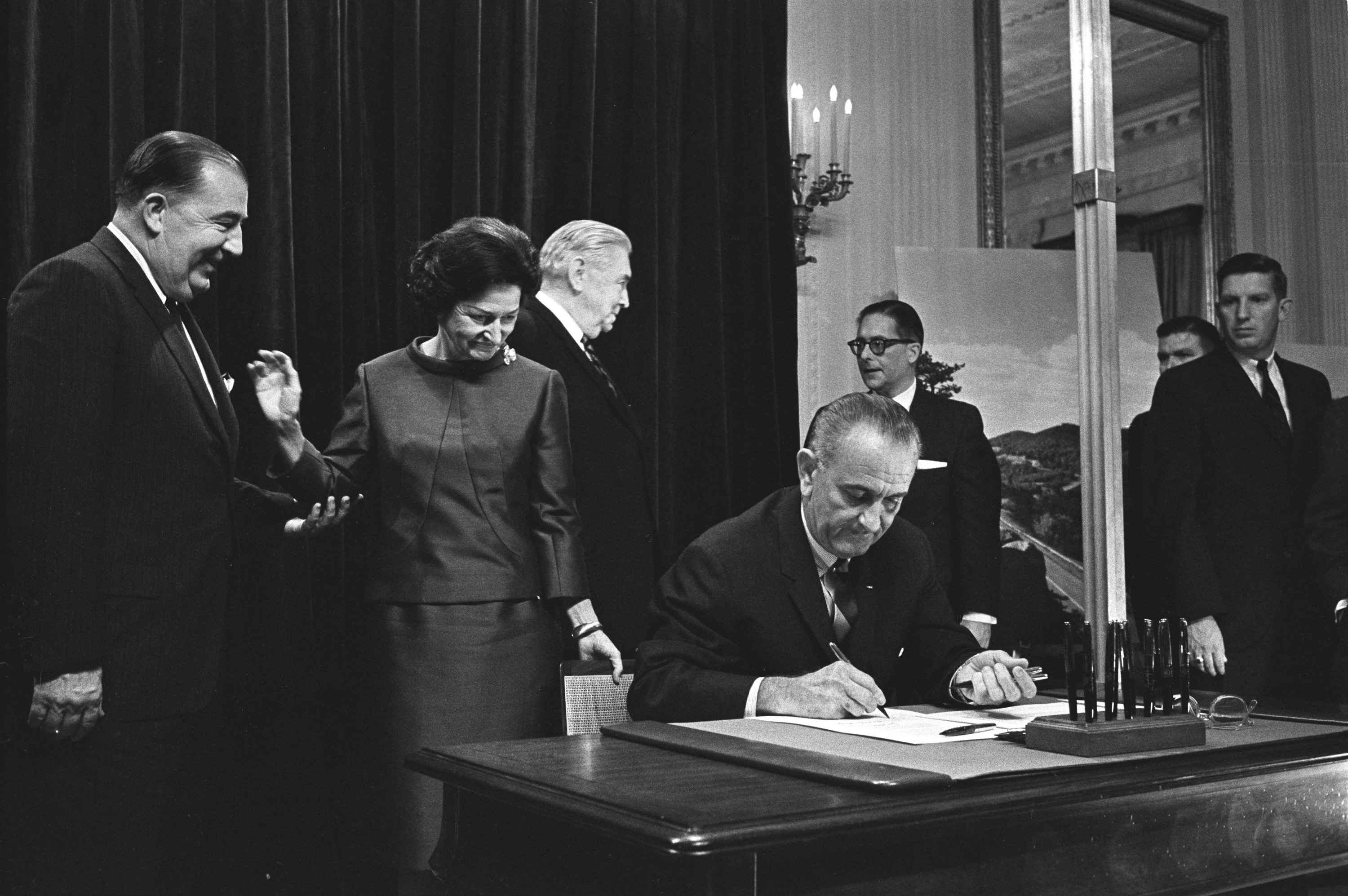 President Lyndon B. Johnson signs the Highway Beautification Bill. Others including Sen. Jennings Randolph, Lady Bird Johnson, and Sen. Pat McNamara look on.