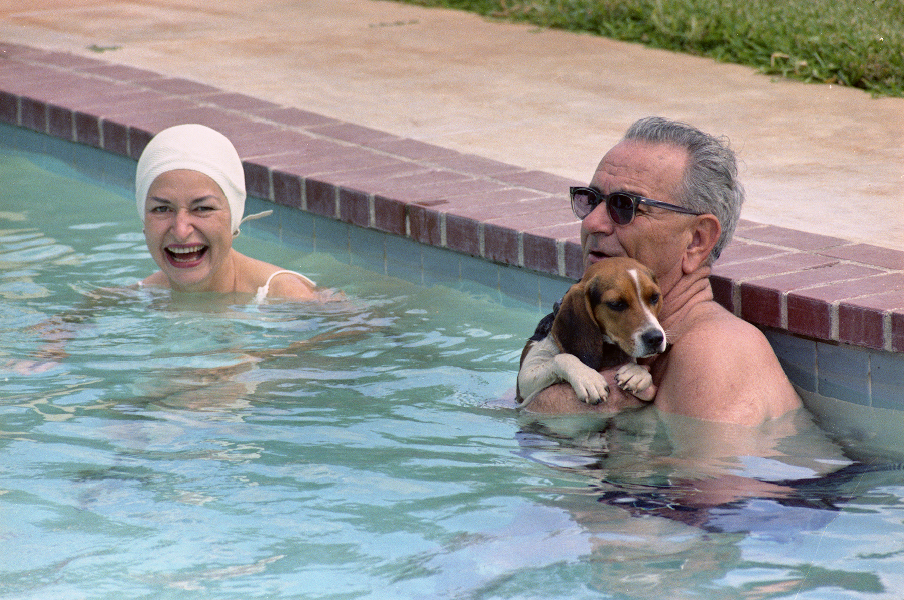 President Johnson, while holding his beagle Freckles, and Lady Bird swim in the LBJ ranch pool.