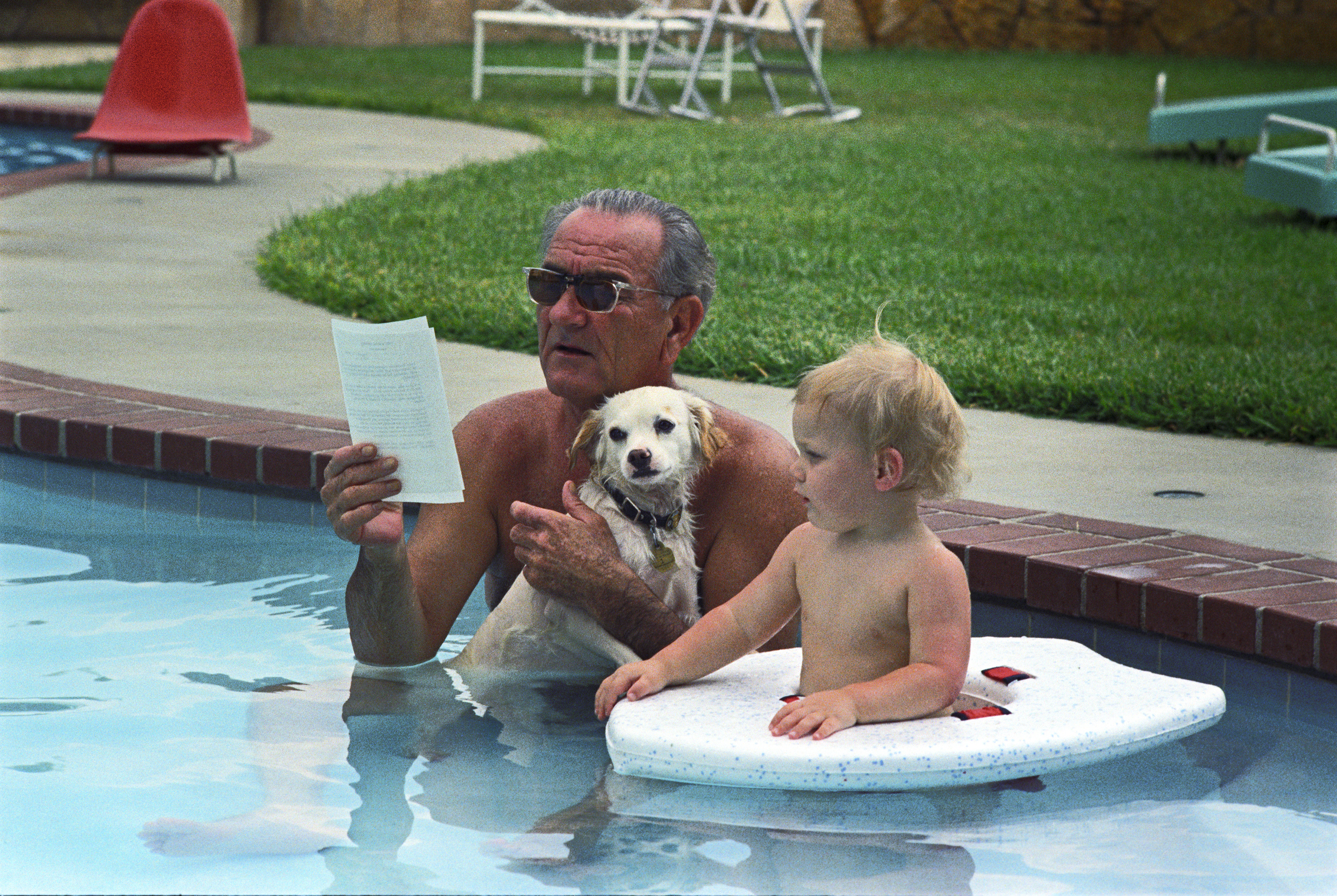 President Johnson, while holding his dog, Yuki, sits in the LBJ ranch pool reading a memo. His grandson, Patrick Lyndon Nugent, is beside him.