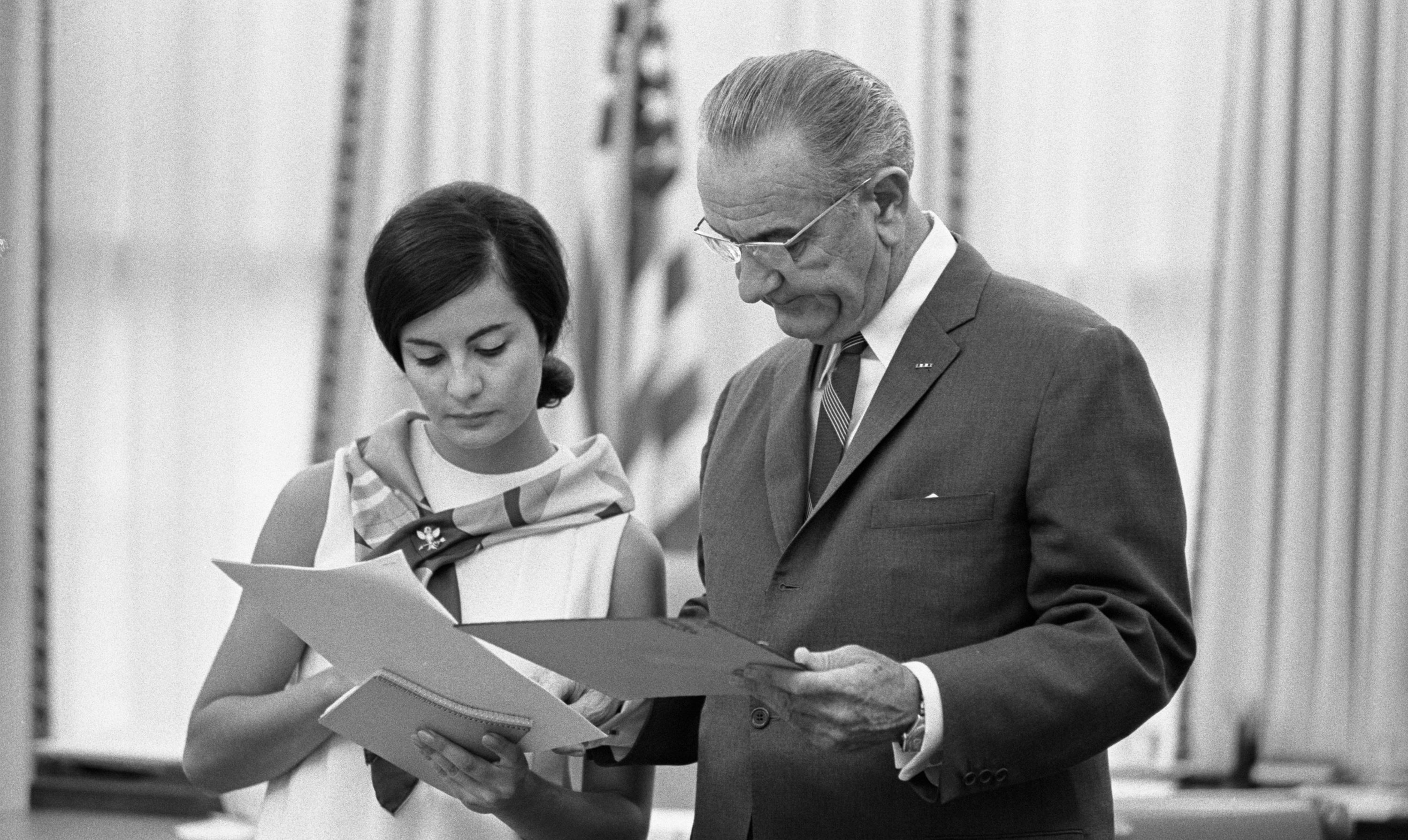 Standing in the Oval Office, President Johnson looks over papers with secretary Phyllis Bonanno.