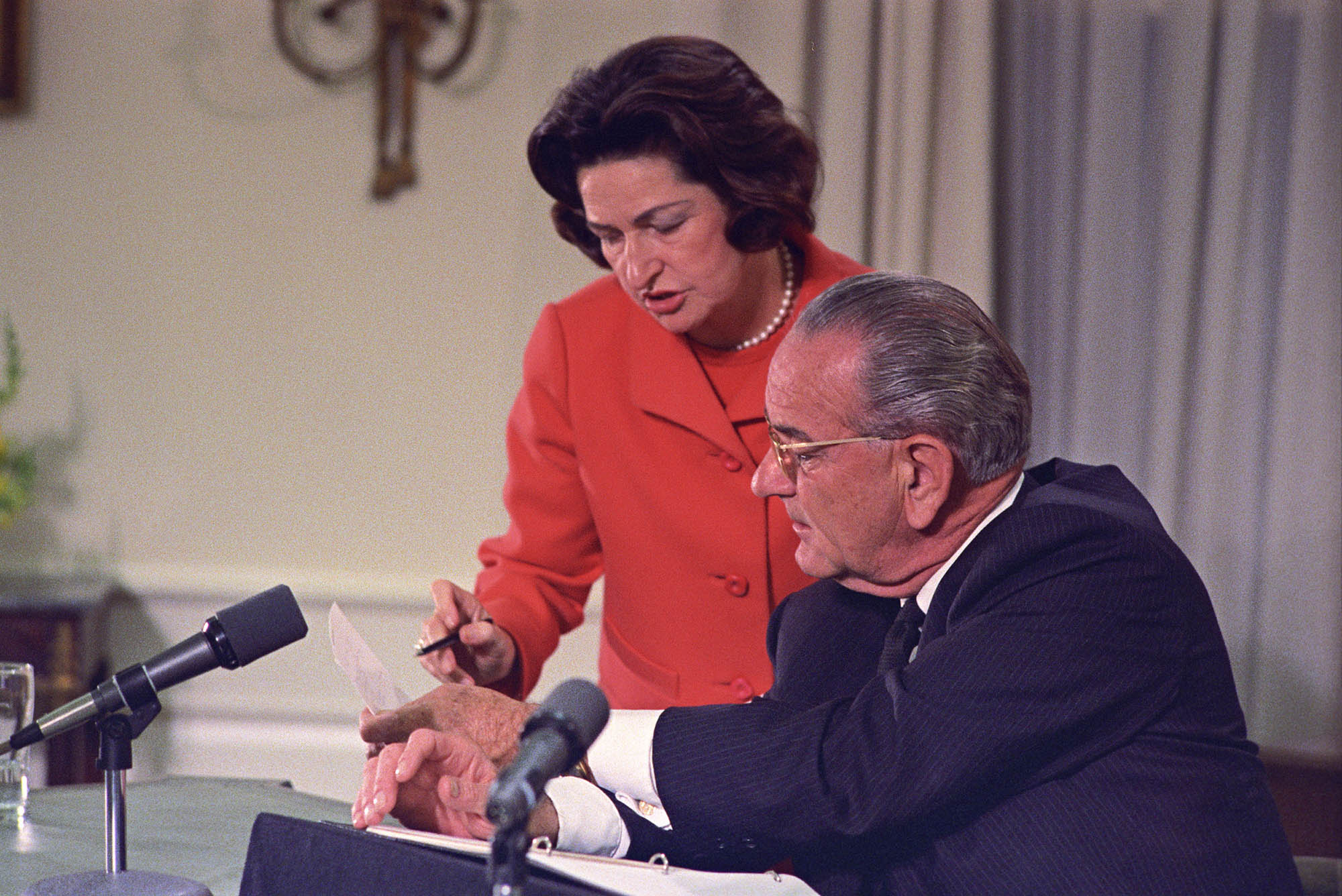 Standing behind President Johnson as he sits at his Oval Office desk, Lady Bird looks over at a paper he is holding.