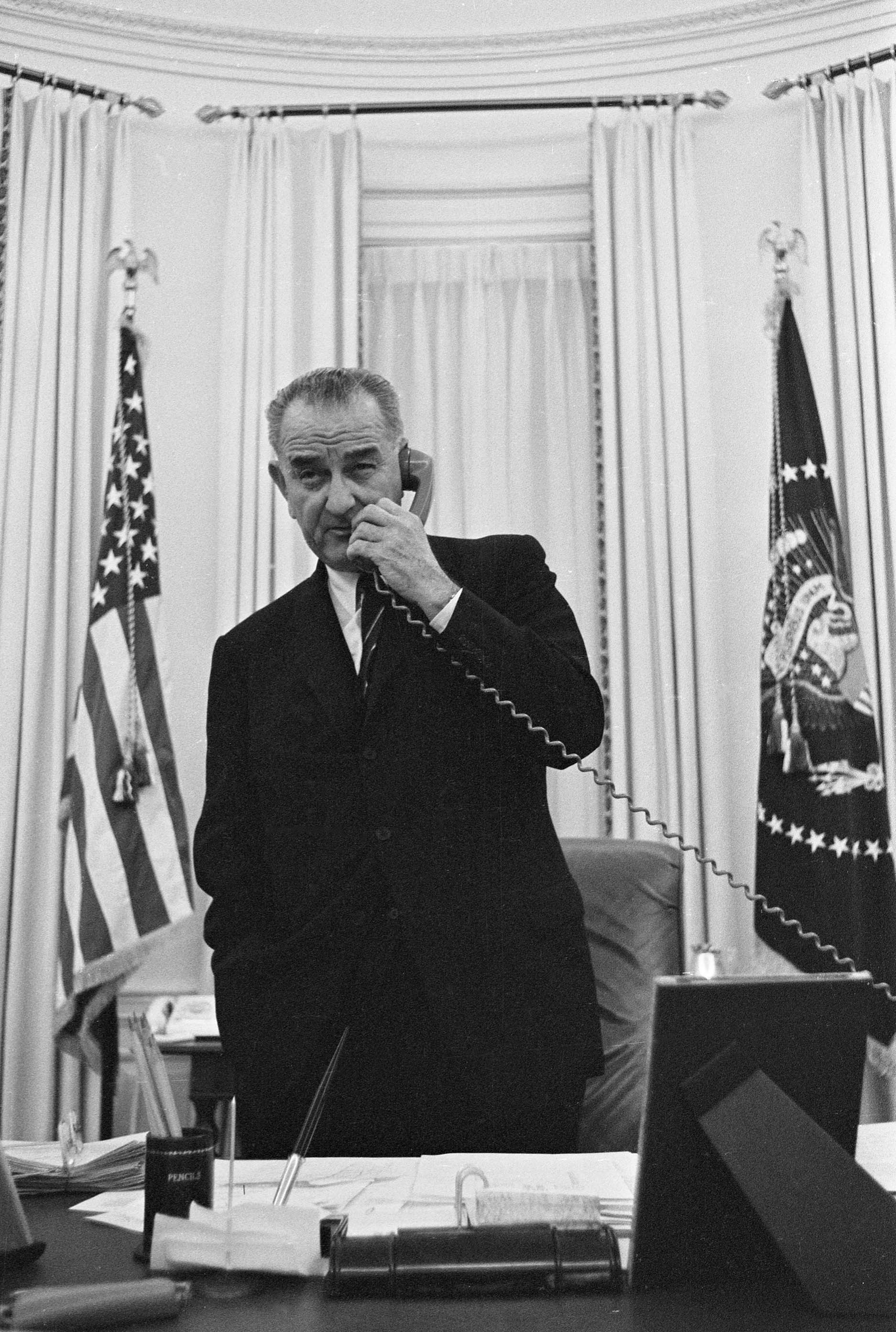 Standing behind his desk in the Oval Office, President Johnson speaks on the telephone.