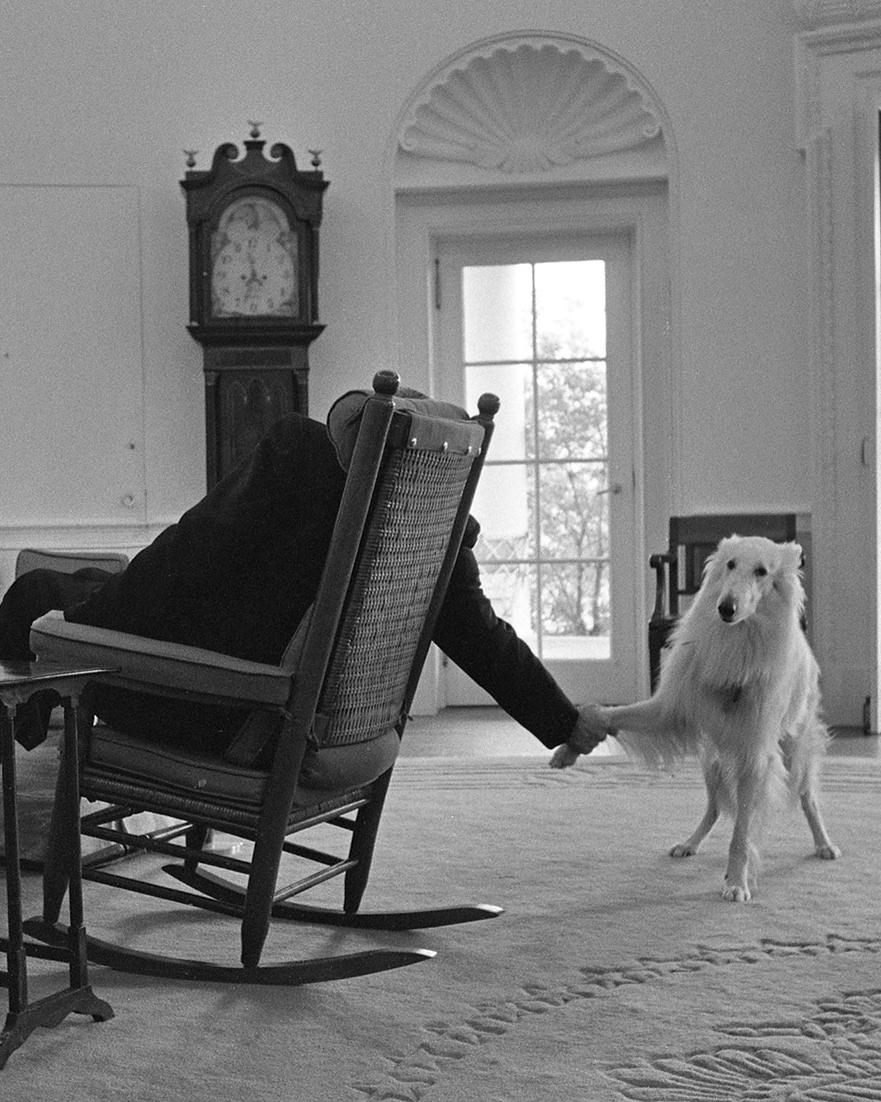 Seen from behind, President Johnson holds Blanco's paw while sitting in his rocking chair in the Oval Office.