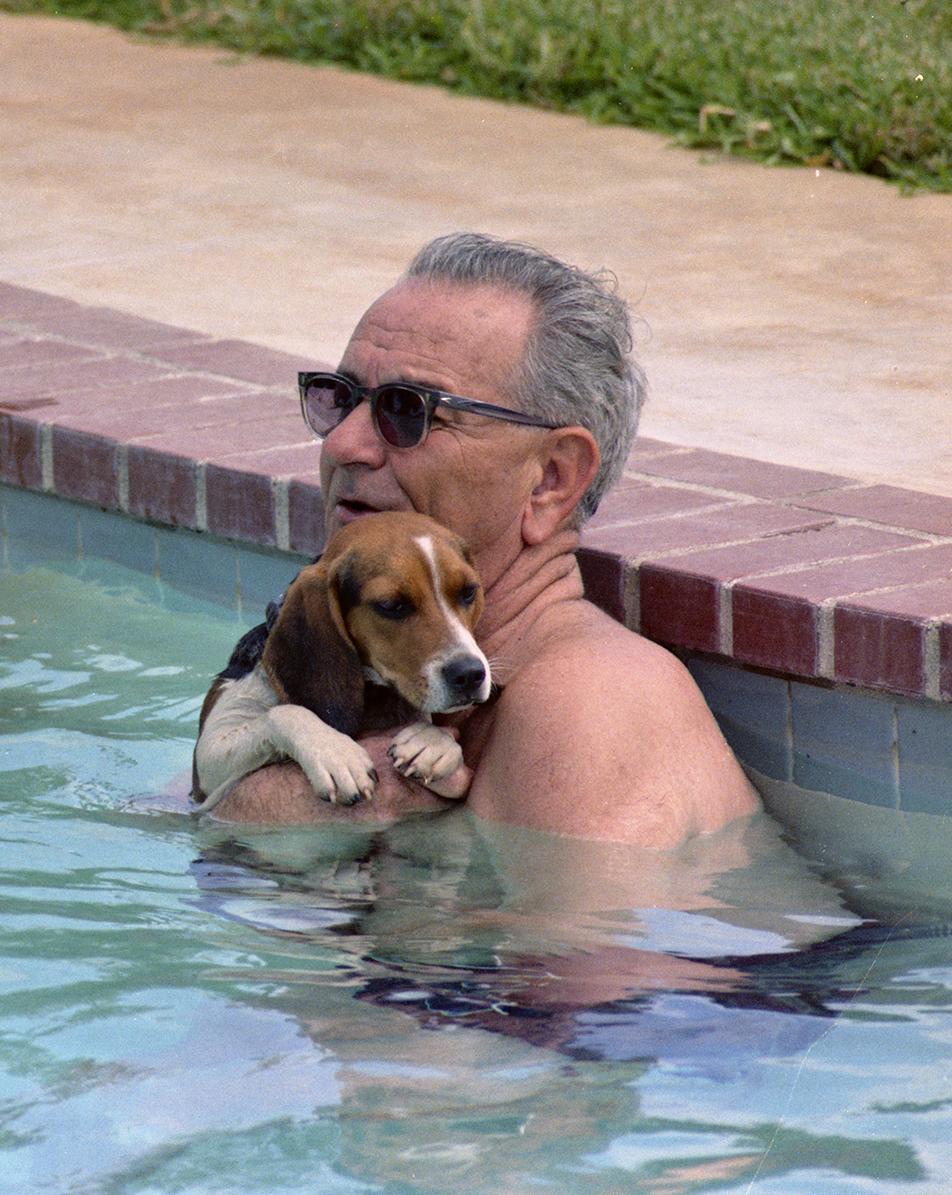 President Johnson, while holding his beagle Freckles, and Lady Bird swim in the LBJ ranch pool.