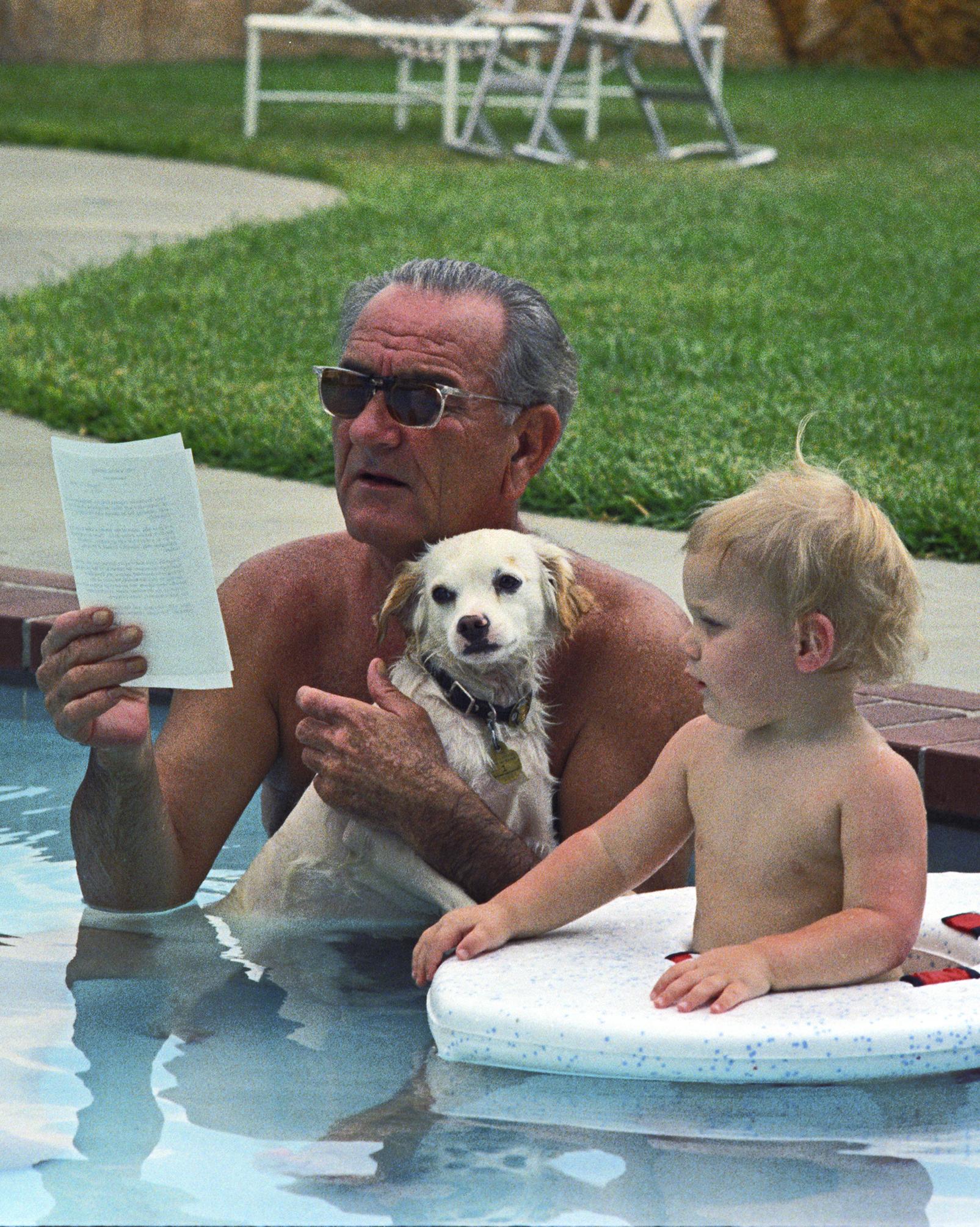 President Johnson, while holding his dog, Yuki, sits in the LBJ ranch pool reading a memo. His grandson, Patrick Lyndon Nugent, is beside him.