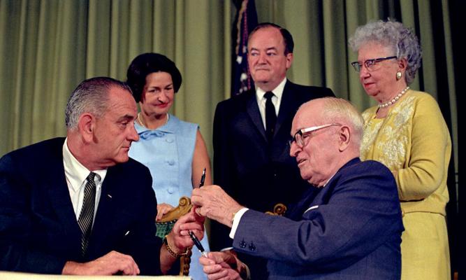 President Lyndon B. Johnson hands President Harry S. Truman a pen as Lady Bird Johnson, Vice President Hubert Humphrey, and Bess Truman look on. LBJ Library photo by Unknown #34896-10A.