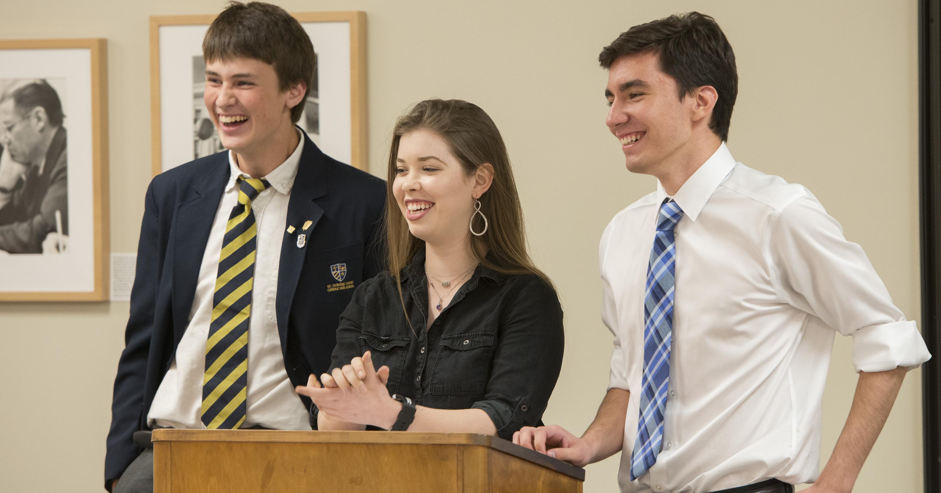Three students, two male and one female, laugh while standing at the podium presenting their findings during an immersive classroom experience at the LBJ Library.